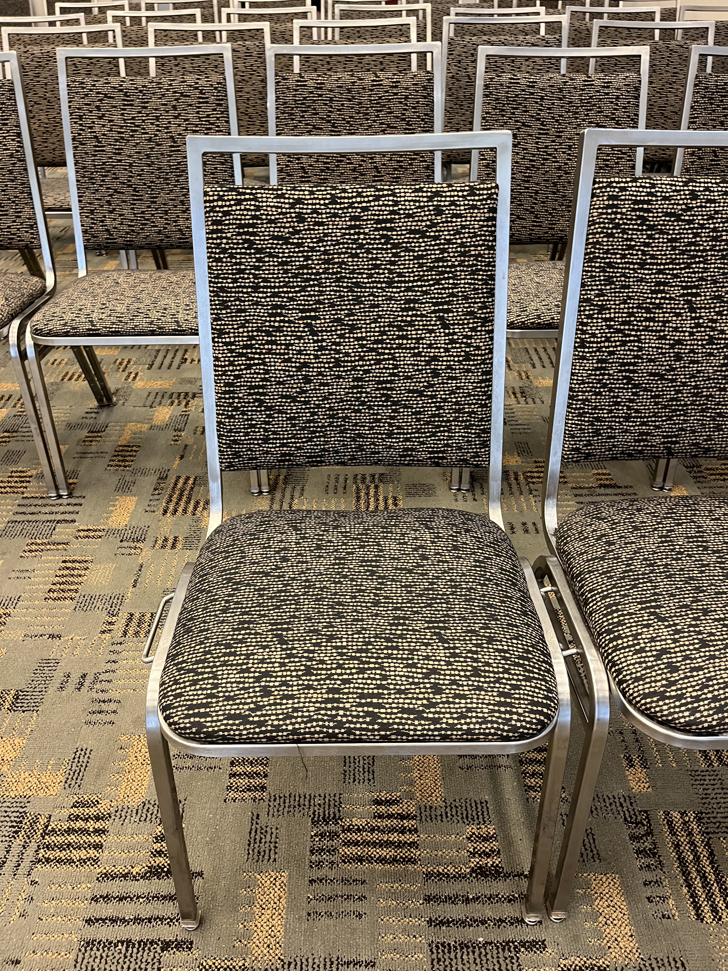 A photo of a metal frame chair with seat and back padding in a gray and black pattern. The chair is in a row and linked to the chair beside it.