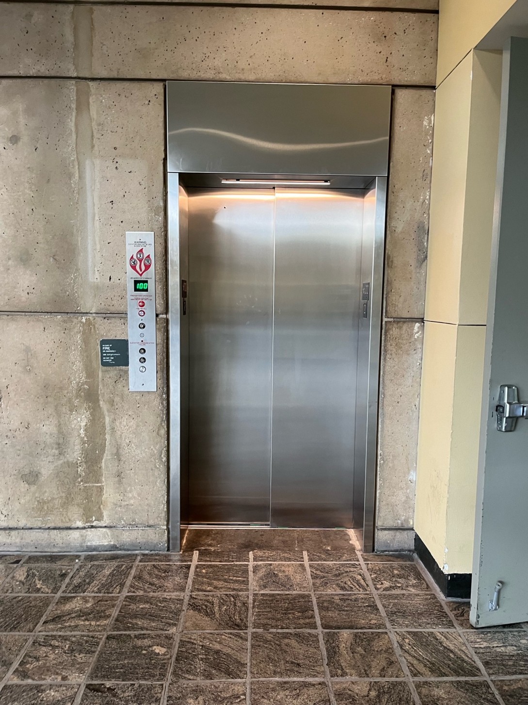 A photo of the level 100 Charles Street Lobby elevator with the panel of buttons on the left. The elevator is silver metal and the surrounding walls are beige concrete with the same gray and beige tile floor as the rest of the Charles Street Lobby.